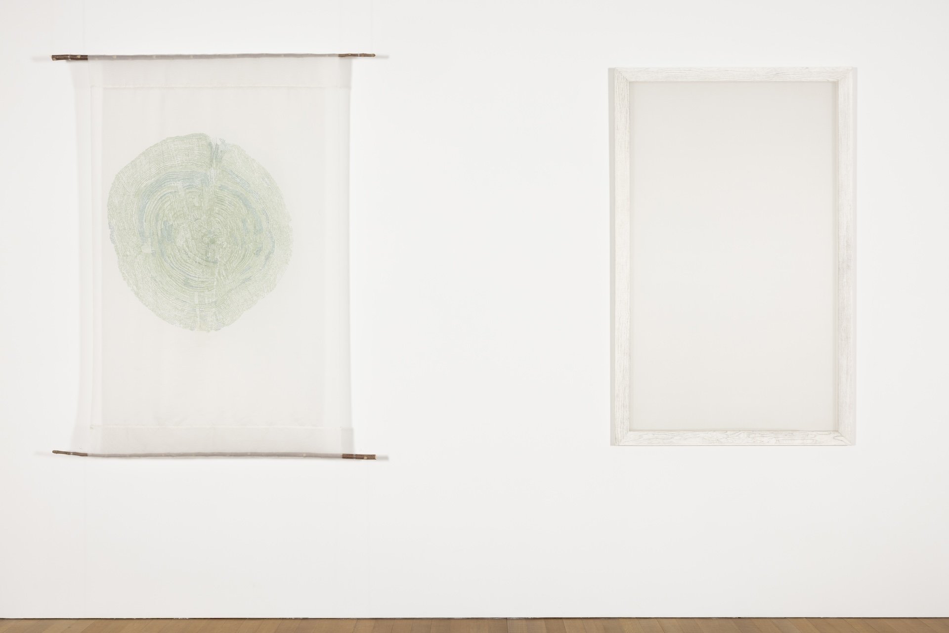 Two rectangular paintings hang on the white wall. The left work is a piece of translucent white silk painted with the circular cross-section of a green tree trunk and with horizontal wooden rods sewn at the top and bottom. The right work is a piece of translucent white silk mounted on a white wooden frame.
